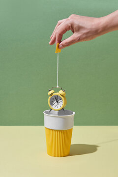 Woman's hand holds alarm clock as a teabag above cup.
