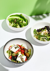 Greek salad, cheese and beet salad and cucumbers with chuka seaweed. Salad bowl on white table with green wall. Sunlight with harsh shadow of green tropic leaf.