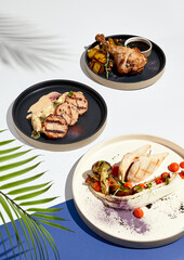 Various meat poultry dishes on white and blue table. Chicken and turkey poultry food plate. Restaurant plate in modern food style. Sunlight with harsh shadow of palm leaves.