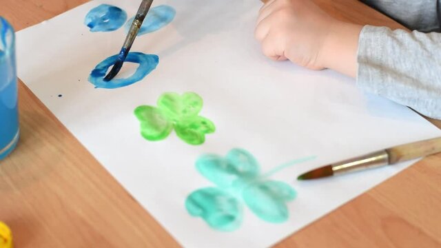 A tooddler drawing a leaves of clover with paintbrush and watercolors on a white sheet of paper.Hands close up.Concept of St.Patricks Day.