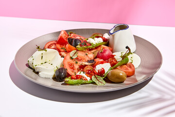 Tomato, olives and mozzarella salad . Restaurant appetizer plate on white table with pink wall. Day sunlight with hard shadow of fern palm leaves. Summer or spring restaurant food concept