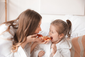 Young woman with her little daughter eating pizza in bedroom