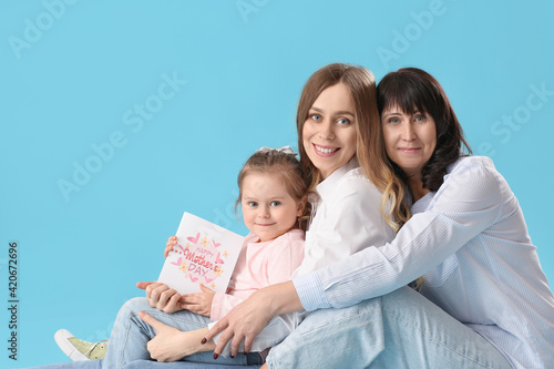 Young woman, her little daughter and mother with greeting card on color background. Mother's Day celebration