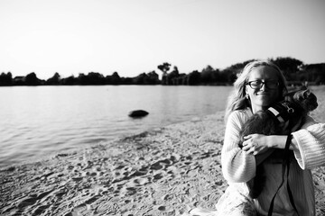 Young blonde girl with glasses holds a cat breed Scottish Straight. Walk with pet near the lake, river at sunset on the beach