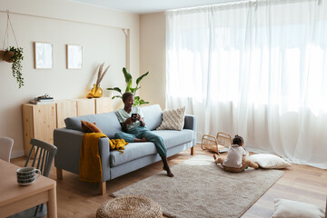 Mother with smartphone resting on sofa near little kid
