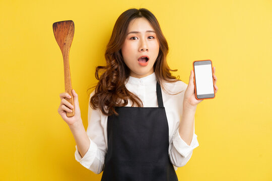 Beautiful housewife holding rice spoon and phone with happy surprise isolated on yellow background