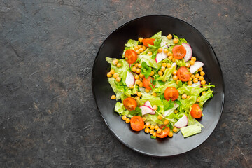 A healthy salad of green lettuce, radishes, tomato with boiled chickpeas on a black plate on a dark concrete background. Served with natural yoghurt sauce. Salad recipes.