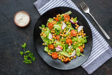 A healthy salad of green lettuce, radishes, tomato with boiled chickpeas on a black plate on a dark concrete background. Served with natural yoghurt sauce. Salad recipes.