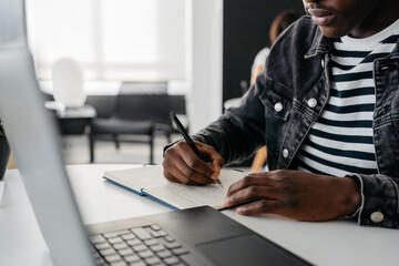 Anonymous young black man remote learning viewing a video on his laptop and taking notes of the explanations