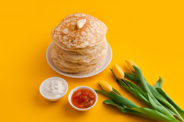 Staple of yeast pancakes with butter and bouquet of tulips, sause bowls with toppings sour cream and jam. Spring holiday Traditional Russian pancake week. isolated on yellow background
