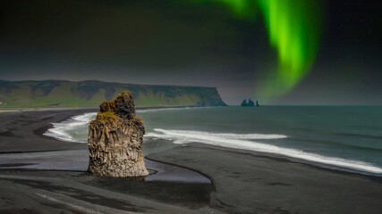The Arnardrangur basalt sea stack on its black volcanic beach at low tide, with the northern lights in the night sky, looking toward the Reynisfjara rocks from Dyrhólaey in southern Iceland.