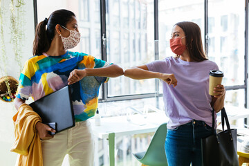Young multiethnic women in masks bumping elbows in workspace during coronavirus pandemic