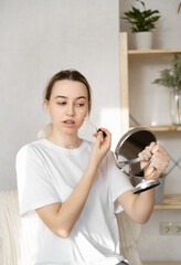 a young teenage woman in a white T-shirt masks pimples on her face with a foundation in front of a mirror. Care for sick skin of the face. Red spots on the skin. Skin problems in transition age