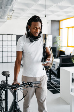 Black young man checking his smartphone while walking in a coworking carrying a bike