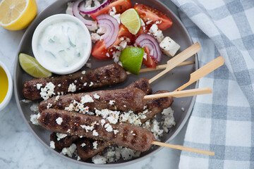 Grey plate with bbq greek kofta kabobs, tzatziki sauce, feta cheese and salad, view from above,...