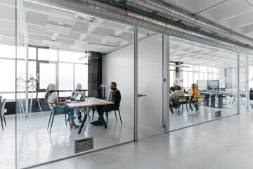 Two different multiethnic groups having a meeting in two glass cubicles of a modern coworking