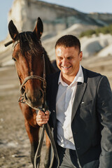 Fototapeta premium Young man dressed in a classic black suit standing with a horse near the mountain