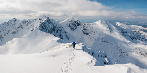 Panoramic view and active man ski touring at mountains background at sunny winter day. Ski mountaineer with blue jacket walking up along a steep snowy ridge with the skis in the backpack