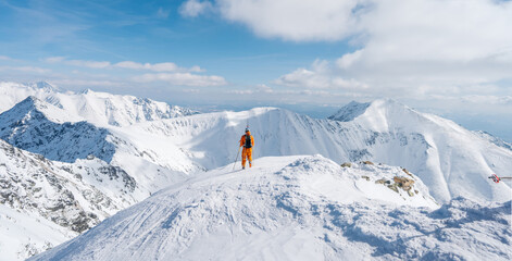 Ski mountaineer walking up along a steep snowy ridge with the skis in the backpack. Climber in a orange jacket climbs a mountain against a blue sky. Adventure concept. Panoramic view 