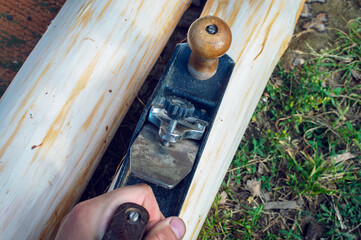 The hand of a young man holds an old manual plane and planes logs outdoors among the green lawn. Top view from the first person