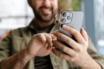 Unshaven happy man smiling and using mobile phone while sitting in cafe