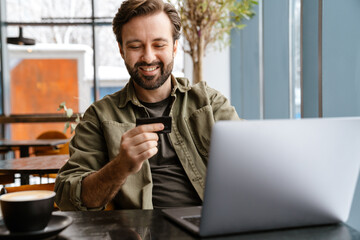 Unshaven happy man using laptop and credit card while sitting in cafe