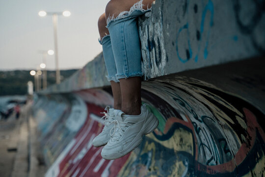 Detail of legs and shoes hanging from wall Young stylish woman sitting on top of wall with graffity with cool attitude