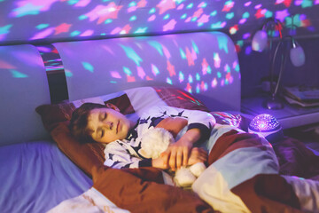 Little kid boy sleeping in bed with colorful lamp. School child dreaming and holding plush toy. Kid...