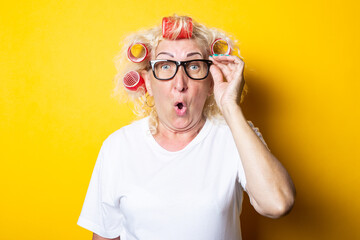 Surprised shock old woman with curlers in glasses looks to the side on a yellow background