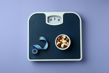 Scales with measuring tape and bowl of nuts on violet background