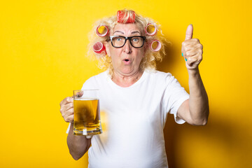 Surprised old woman with hair curlers with a glass of beer showing thumb up hand gesture on yellow background