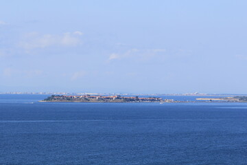 In the middle of the blue sea there is a peninsula with low old buildings and the other shore on the horizon