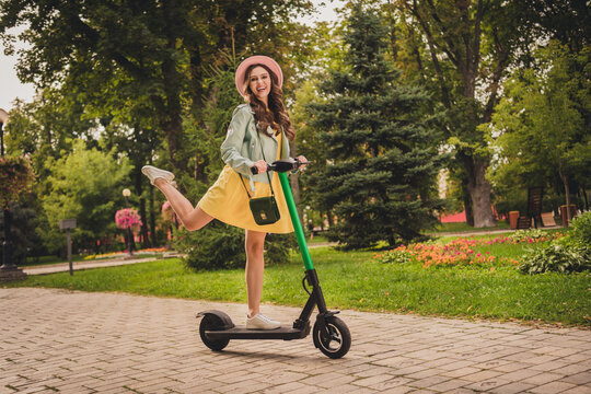 Photo of cute carefree young woman dressed teal jacket glasses cap riding kick scooter rising leg outdoors city park