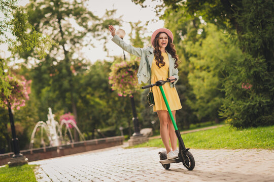 Photo of adorable young lady wear turquoise jacket spectacles headwear handbag waving arm driving scooter outside city park