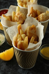 Paper cups with fried fish and chips, and sauces on black smokey background