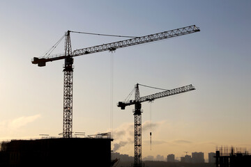Silhouettes of construction cranes and unfinished residential building against the sunrise above the city. Housing construction, apartment block