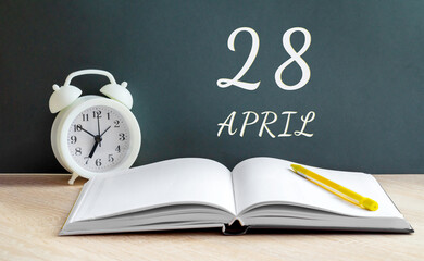 April 28. 28-th day of the month, calendar date.A white alarm clock, an open notebook with blank pages, and a yellow pencil lie on the table.Spring month, day of the year concept