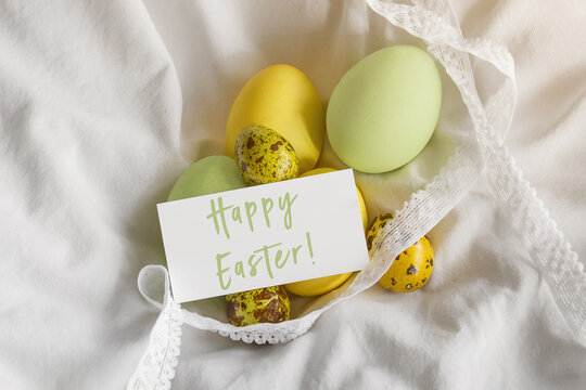 painted easter yellow chicken and quail eggs, spring mimosa sprig, greeting white card with the inscription happy easter on white cloth