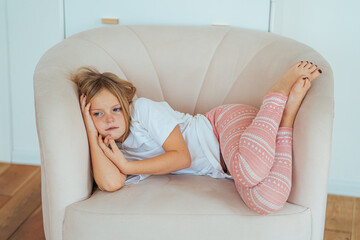 sad girl child in home clothes lies on a beige armchair