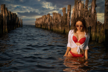 Young beautiful female brunette model pose in her red swimwear lingerie and white shirt over in the water between old pier wood bulks during dramatic sunset