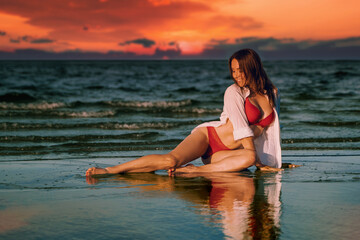 Fototapeta na wymiar Young beautiful female brunette model pose at the beach wet sand in her red swimwear lingerie during dramatic sunset