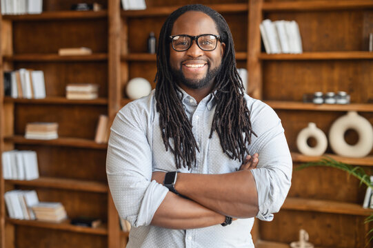 Portrait of a young strong african unshaved man with dreadlocks in glasses stands with arms crossed on his chest, looks at the camera, smiles, against the background of shelves with books, posing