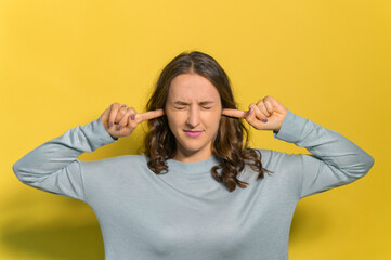 A young woman with closed eyes is plugging ears with fingers, irritated from noise and din, closed in myself, isolated on yellow