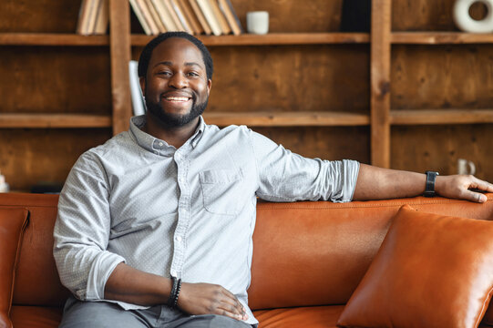 Ethnic black unshaved plump man with dreadlocks sitting on the leather sofa and smiling, in a normal blue shirt and jeans, feeling comfort and relaxation, at an interview, bookcase in the background