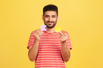 Smiling bearded man in striped t-shirt holding and showing two jigsaw pieces, matching and...