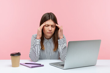 Tired business woman massaging temples feeling unpleasant headache, suffering migraine sitting at workplace with laptop, overwork. Indoor studio shot isolated on pink background