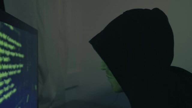 Hacker in front of his computer screen coding and hacking into security systems in dark green room. Hooded guy cyber-attacking an online system breaking into the internet network. Man programming code