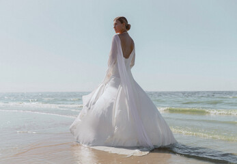 A blonde haired woman standing with her back position on the beach wearing a long bridal gown