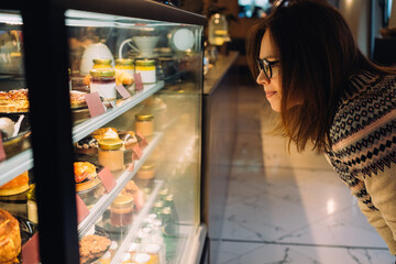 Young woman looking on the showcase with different desserts in coffee shop
