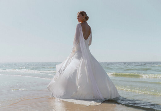 A blonde haired woman standing with her back position on the beach wearing a long bridal gown